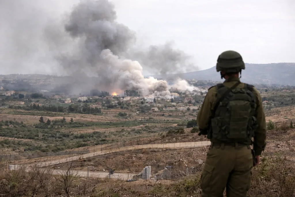 Israeli soldier standing near the border fence while smoke rises over southern Lebanon following reported airstrikes