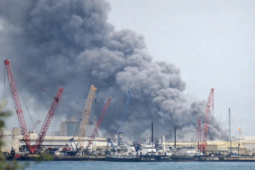 Thick smoke rising above a port area in Manama, Bahrain, with cranes and docked vessels visible in the foreground